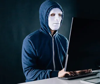 Person in a hoodie and white mask typing on a computer in a dark room, illustrating anonymity or online trolling.