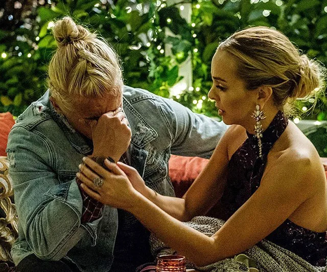 Man with long hair and denim jacket crying, comforted by a woman in a dark dress, surrounded by greenery and fairy lights.