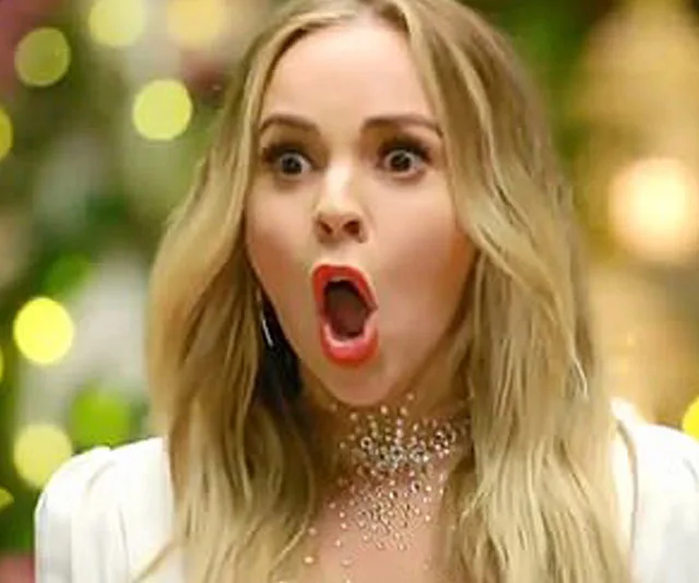 A surprised woman with long blonde hair and red lipstick, mouth agape, against a blurred background with lights.