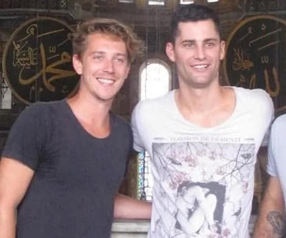 Two men smiling in a historic setting with ornate wall art, both casually dressed.