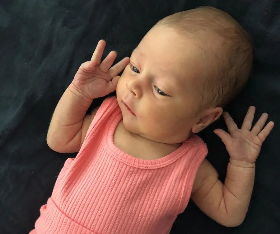 Newborn baby wearing a pink outfit lies on a dark blanket, looking to the side with hands raised near head.