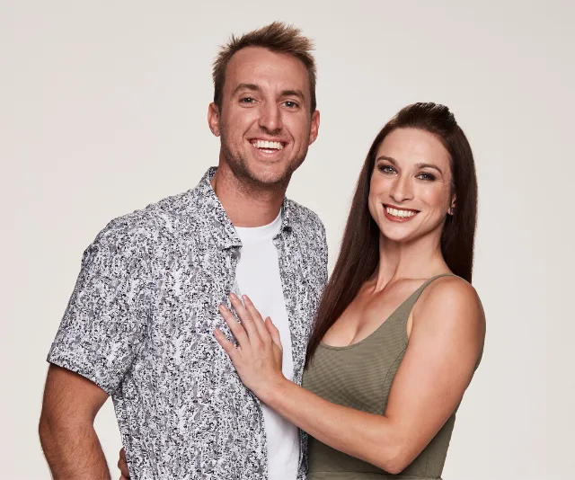 A smiling couple stands together, the woman in a green dress and the man in a patterned shirt against a plain background.