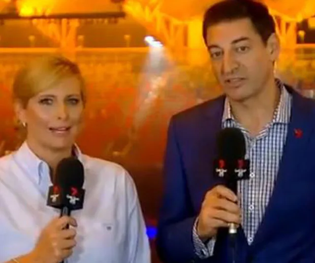 Two TV presenters holding microphones in an arena with warm lighting in the background.