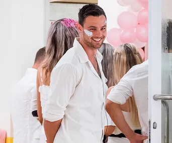 Man in white shirt with face glitter smiles at a party with pink balloons in the background.