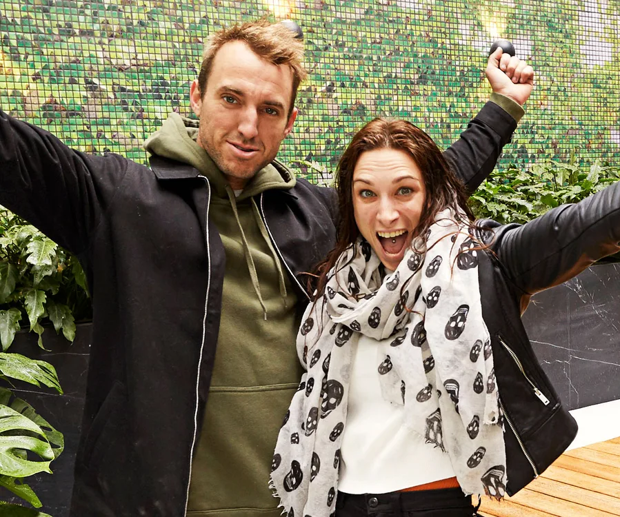 A joyful man and woman with arms raised, celebrating outdoors next to a green wall and plants.