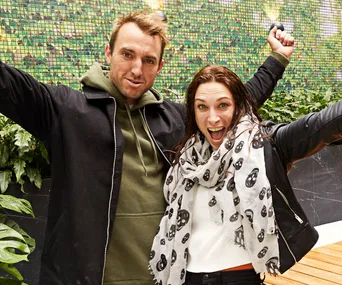 A joyful man and woman with arms raised, celebrating outdoors next to a green wall and plants.