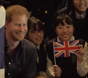 Prince Harry smiling with students, one holding a small UK flag, during a visit to Japan.