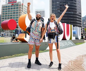 Two people celebrating in front of a colorful "Seoul" sign, both wearing vibrant matching outfits and backpacks.