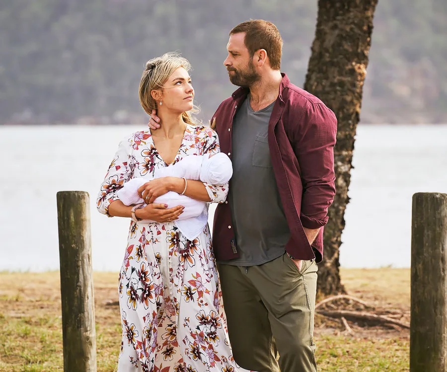Woman holding baby stands beside man outdoors, both look into distance near water and trees.