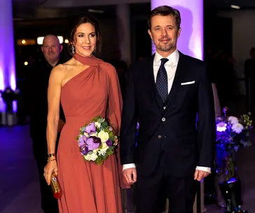 Princess Mary and Prince Frederik at an event; she wears a one-shoulder gown, holds flowers; he wears a dark suit.