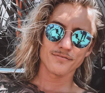 Young man with long hair, wearing reflective sunglasses outdoors, with a slight smile.