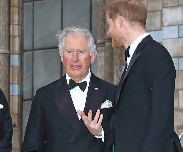 Two men in formal black tuxedos conversing outside a building with stone walls.