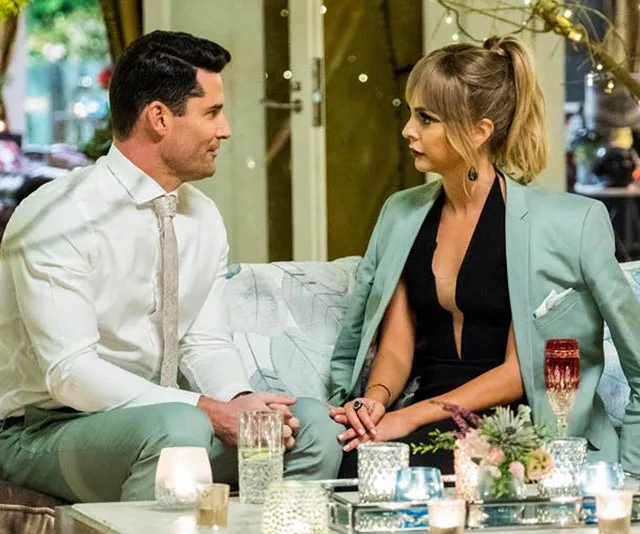 Two people sitting on a couch, dressed formally, having a conversation in a warmly lit room with candles and decorations.