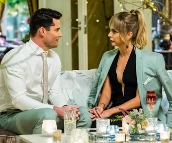 Two people sitting on a couch, dressed formally, having a conversation in a warmly lit room with candles and decorations.