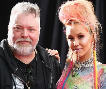 A man with a beard and a woman with colorful hair and a chain accessory, standing together and smiling.