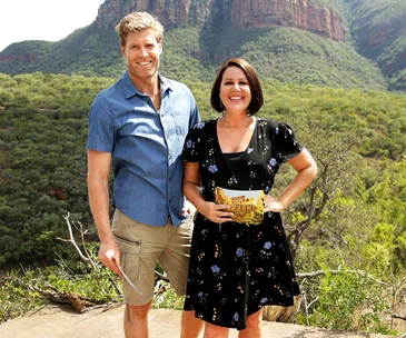 Two hosts smile outdoors in a scenic mountainous area, holding the "I'm a Celebrity" sign.