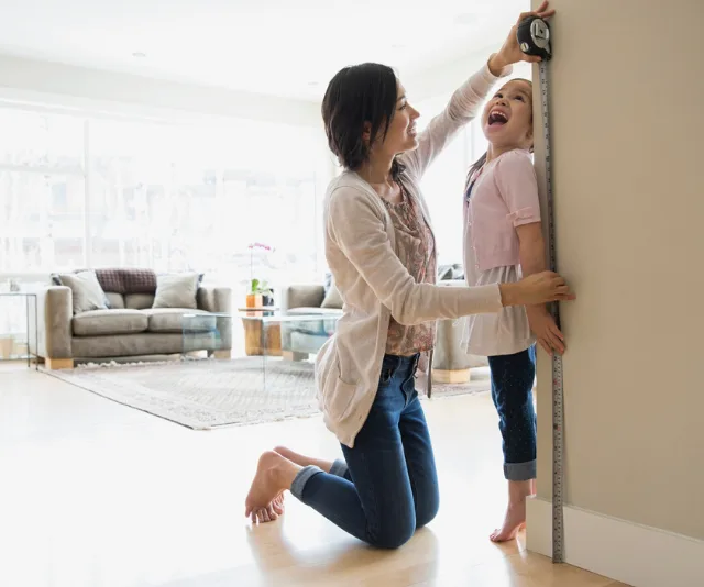 Mother measuring daughter's height with tape measure against a wall, both smiling in a bright living room.