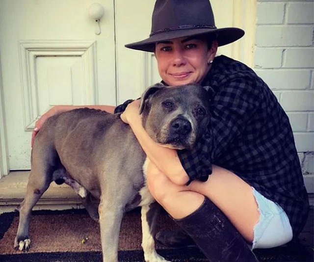 Person in a hat hugging a grey dog in front of a door.