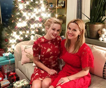 Two women in red dresses sitting on a couch, smiling in front of a decorated Christmas tree with presents.