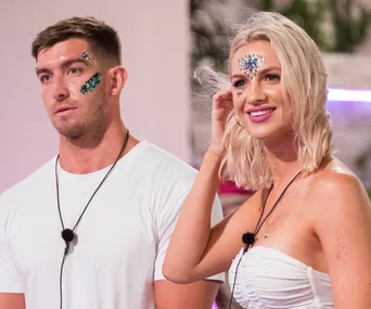 Two individuals with face jewels and mics, standing together on a reality show set.