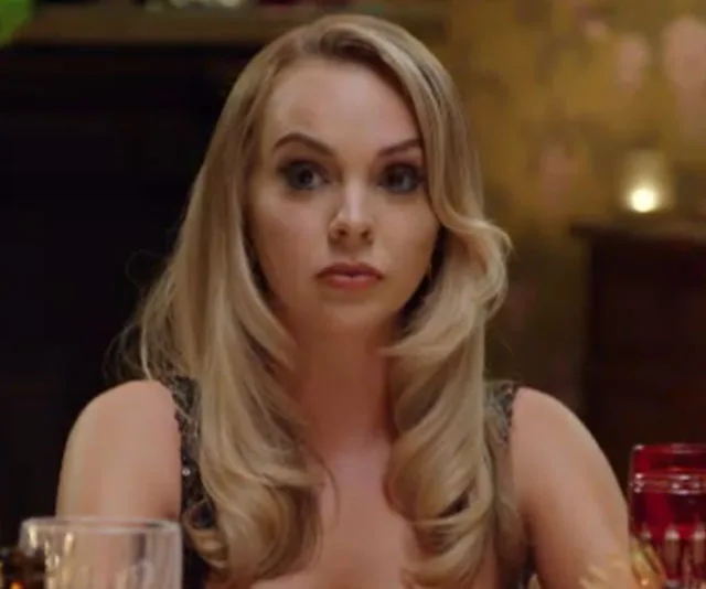 A woman with long blonde hair sitting at a formal dinner table, looking slightly pensive.