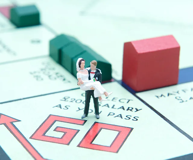 Miniature bride and groom on a Monopoly board's "GO" space with houses around.