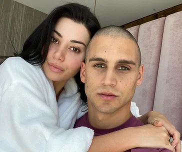 A couple poses closely; the woman in a white robe, the man in a purple shirt, looking directly at the camera.