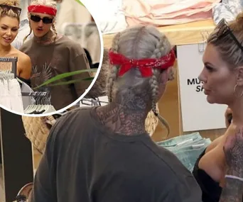 Two individuals are shopping and conversing in a clothing store, one with a red bandana and braids.