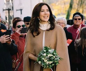 Princess Mary in a beige coat holding a bouquet, smiling at a public event surrounded by onlookers.