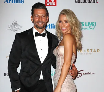 Man in a tuxedo and woman in a strapless dress, smiling on a red carpet event backdrop.