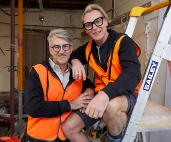 Two men in orange vests sit on a ladder in a construction site, smiling at the camera.