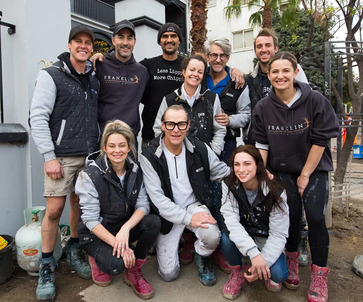 A group of people wearing casual work clothing, posing outdoors, some with paint splatters and branded jackets.