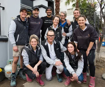 A group of people wearing casual work clothing, posing outdoors, some with paint splatters and branded jackets.