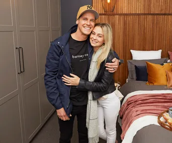 A couple hugging in a well-decorated bedroom with colorful pillows and a tray on the bed.