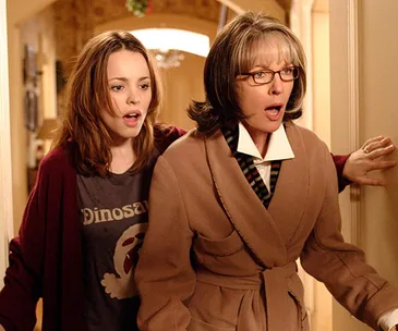 Two surprised women stand in a doorway, one wearing glasses and a coat, the other in a dinosaur T-shirt.