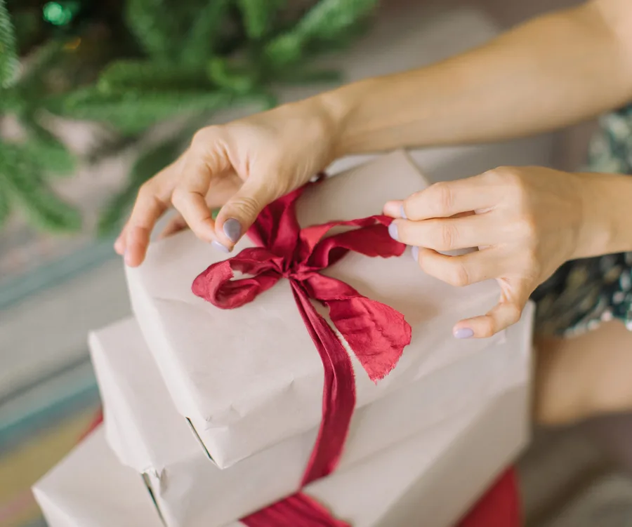 Hands tying a red ribbon on a stack of wrapped gifts under a Christmas tree.