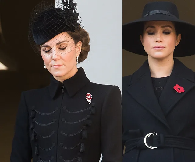 Two women in black coats with poppy pins stand solemnly with eyes closed at a remembrance event.