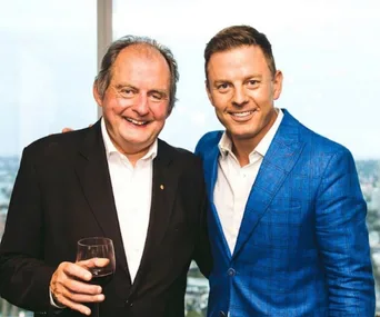 Two men smiling, one in a black suit holding a glass of wine, the other in a blue jacket.