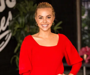 Smiling woman in a red top stands in front of a leafy backdrop.