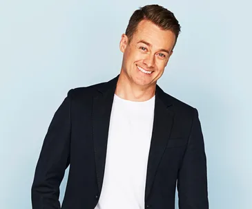 Man in a navy blazer and white shirt smiling against a light blue background.