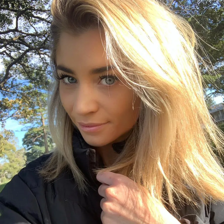 Blonde woman smiles outdoors in sunlight, wearing a black jacket, with trees in background.
