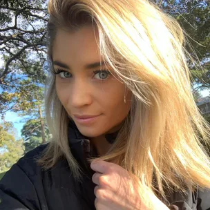Blonde woman smiles outdoors in sunlight, wearing a black jacket, with trees in background.