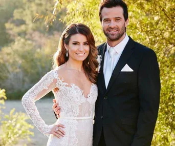 A couple posing outdoors; the woman in a lace wedding dress and the man in a black suit with a white tie.