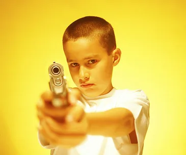 A boy in a white shirt points a handgun directly at the camera against a yellow background.
