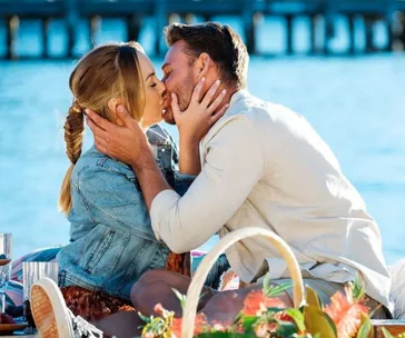 A couple kissing by the water on a sunny day, with a picnic basket in the foreground.