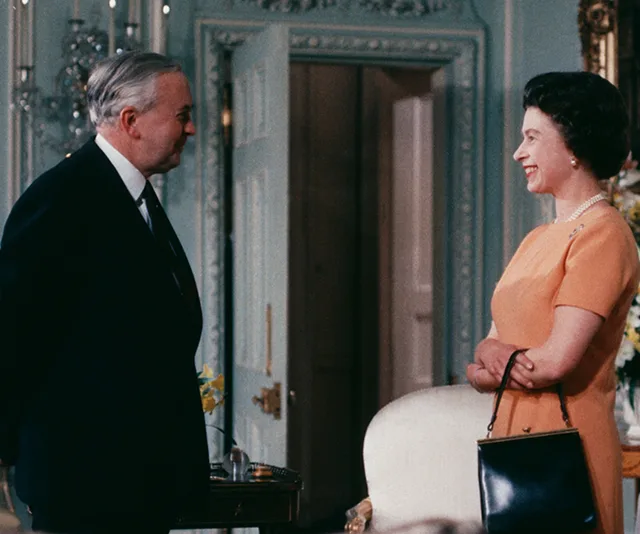 "Two people, a man in a suit and a woman in an orange dress, smiling in a formal room."