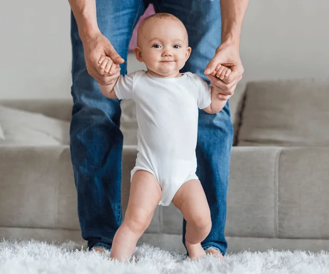 A baby in a white onesie stands on a carpet, holding an adult's hands for support.