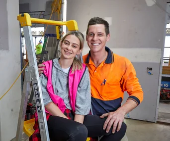A smiling couple in casual work attire inside a construction site, one sitting on a ladder, the other standing beside.