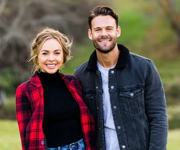 A smiling woman in a red plaid coat stands next to a man in a black jacket outdoors.