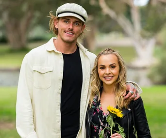 A man and woman smiling outdoors, standing close with trees in the background, the woman holds a sunflower.